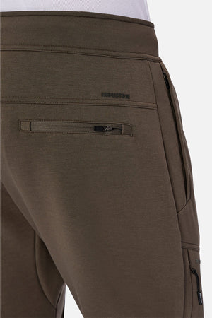 Close-up of a person wearing olive green Industrie The Tech Armoured Track Pant joggers featuring zippered back and combat pockets, with "INDUSTRIE" embroidered above the back pocket.