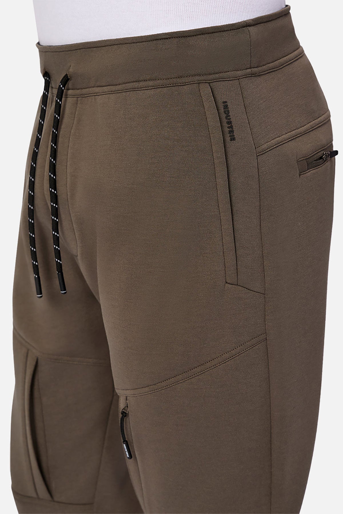 Close-up of a person wearing Industrie The Tech Armoured Track Pant in olive green, featuring technical fabric, black and white drawstrings, zippered combat pockets, visible stitching, paired with a white shirt.