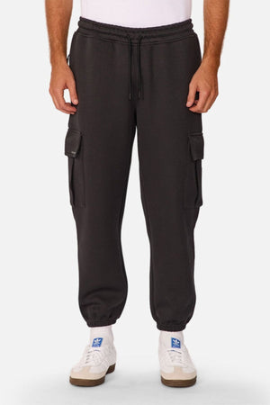 A person wearing Industrie The Tech Combat Trackpant in black with side pockets, white socks, and white sneakers stands against a plain light background. Its customizable fit combines classic style with modern athletic versatility.