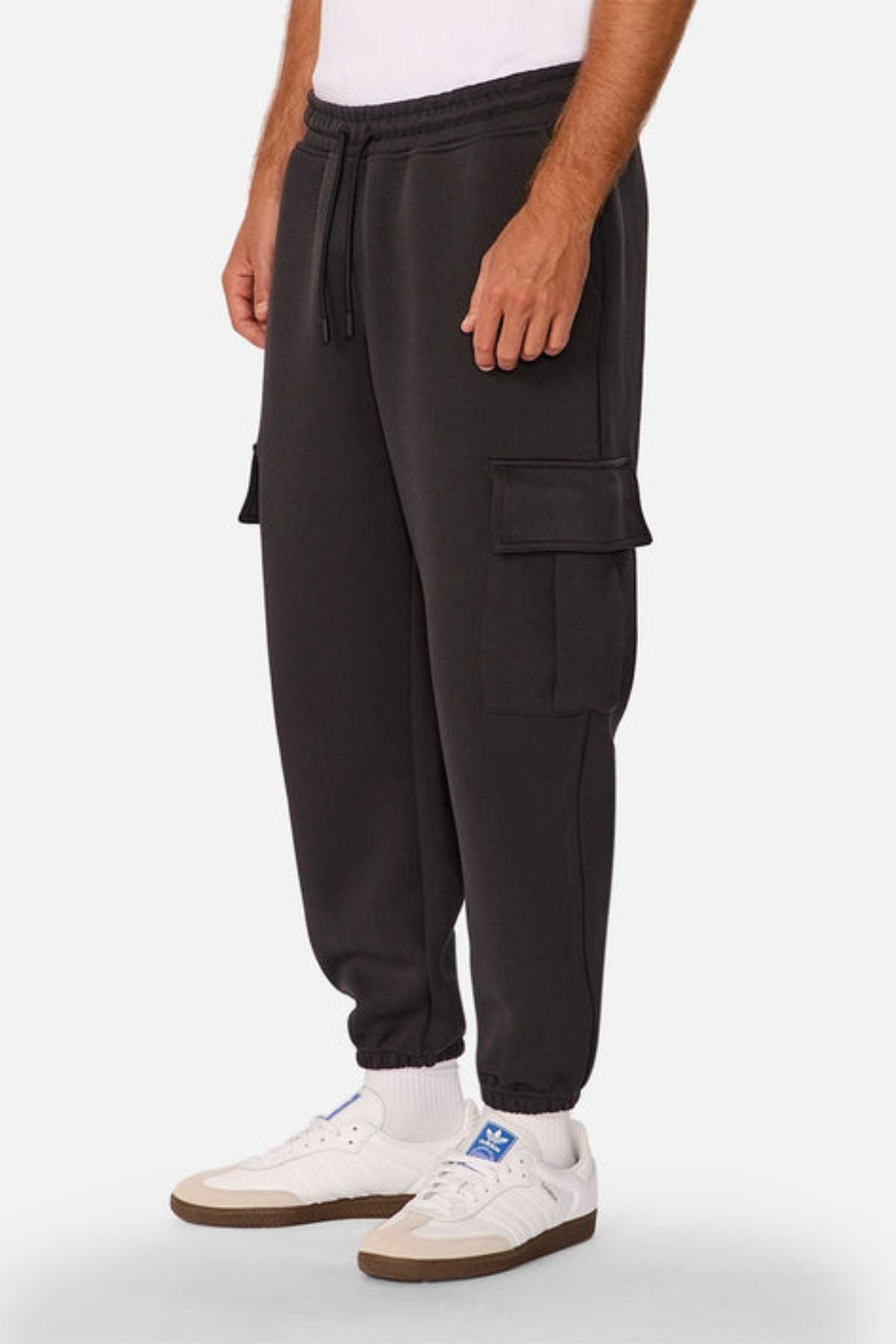 A person wears Industrie The Tech Combat Trackpant cargo sweatpants with side pockets and a customizable fit, paired with white socks and blue-branded Adidas sneakers, standing against a plain background for an athletic style.