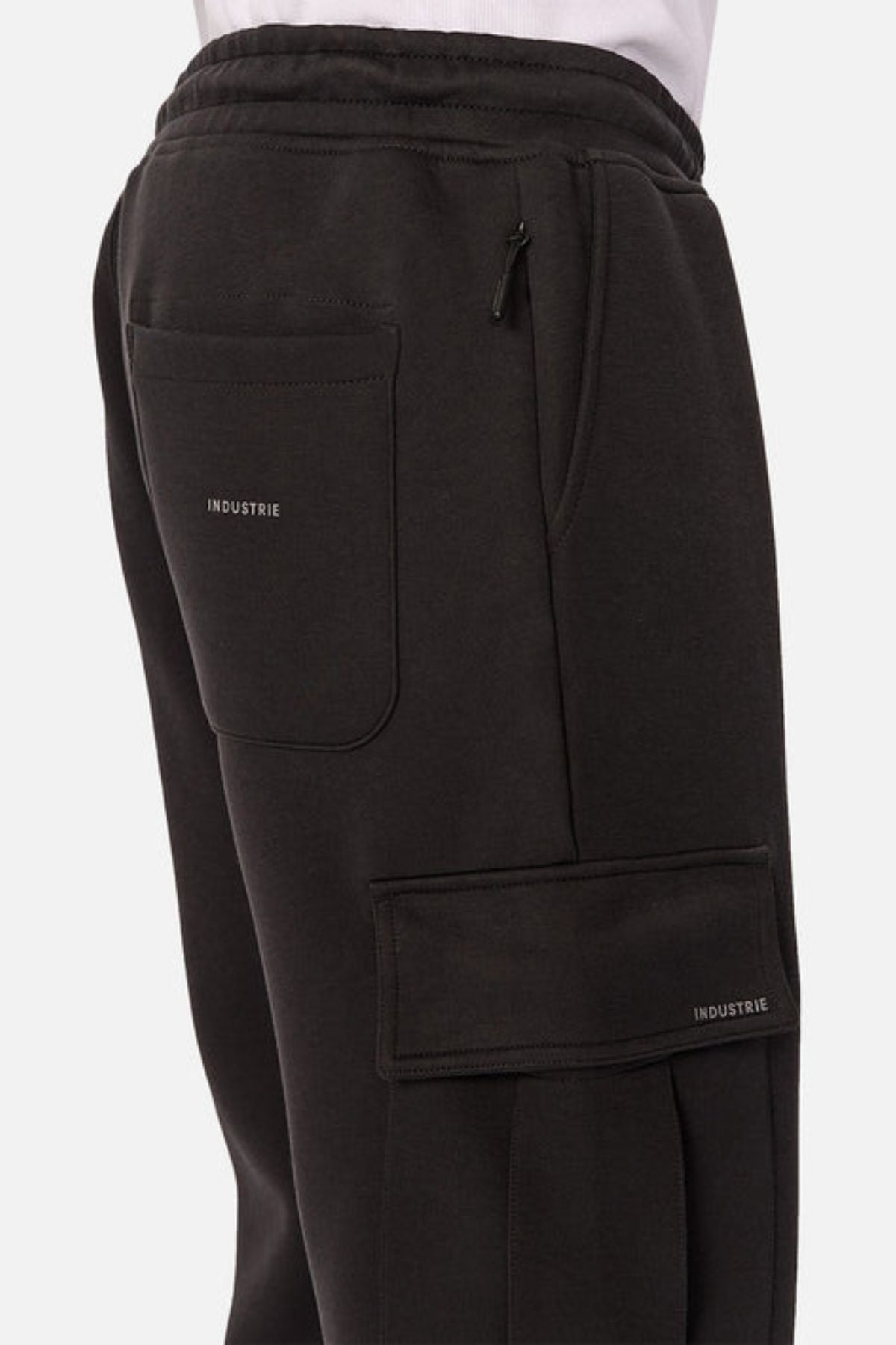 A close-up side view shows someone wearing Industrie The Tech Combat Trackpant in black, featuring multiple pockets (one zippered, one labeled “INDUSTRIE”), paired with a white shirt for a customizable athletic look.