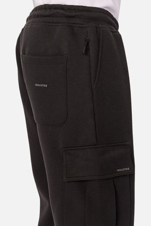 A close-up side view shows someone wearing Industrie The Tech Combat Trackpant in black, featuring multiple pockets (one zippered, one labeled “INDUSTRIE”), paired with a white shirt for a customizable athletic look.