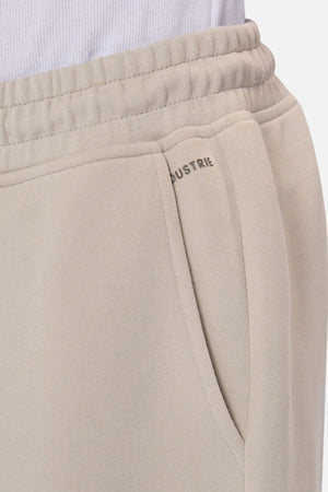 Close-up of someone in Industrie The Tech Combat Trackpant, light beige with elastic waistband and side pocket. The "INDUSTRIE" logo is partly visible near the pocket, styled with a white shirt for an effortless athletic look.