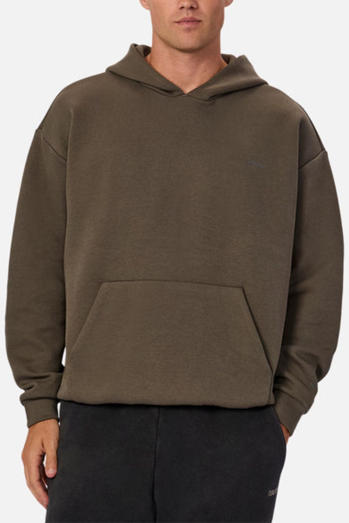 A person wears the oversized Industrie The Tech Del Sur Hood in plain olive green, featuring a front pocket, hood, and long sleeves made from advanced athletic fabrications, paired with black pants against a light background.
