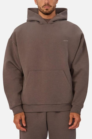 A man wears the Industrie The Tech Del Sur Hood, a loose-fitting brown sweatshirt with matching sweatpants made from advanced athletic fabrics, featuring the INDUSTRIE logo, standing against a plain white background.