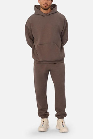 A man wears the Industrie The Tech Del Sur Hood in loose-fitting brown with matching joggers and off-white sneakers, standing hands behind his back against a plain, light background.