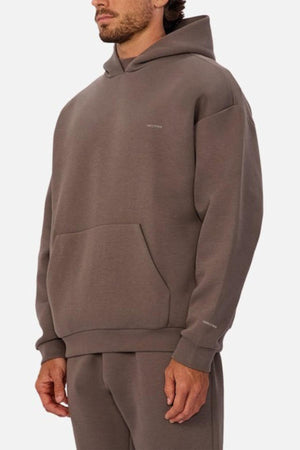 A man wears the Industrie The Tech Del Sur Hood in brown with matching sweatpants, featuring advanced athletic fabric, a front pocket, minimal chest text, and the INDUSTRIE logo. He stands against a plain white background.