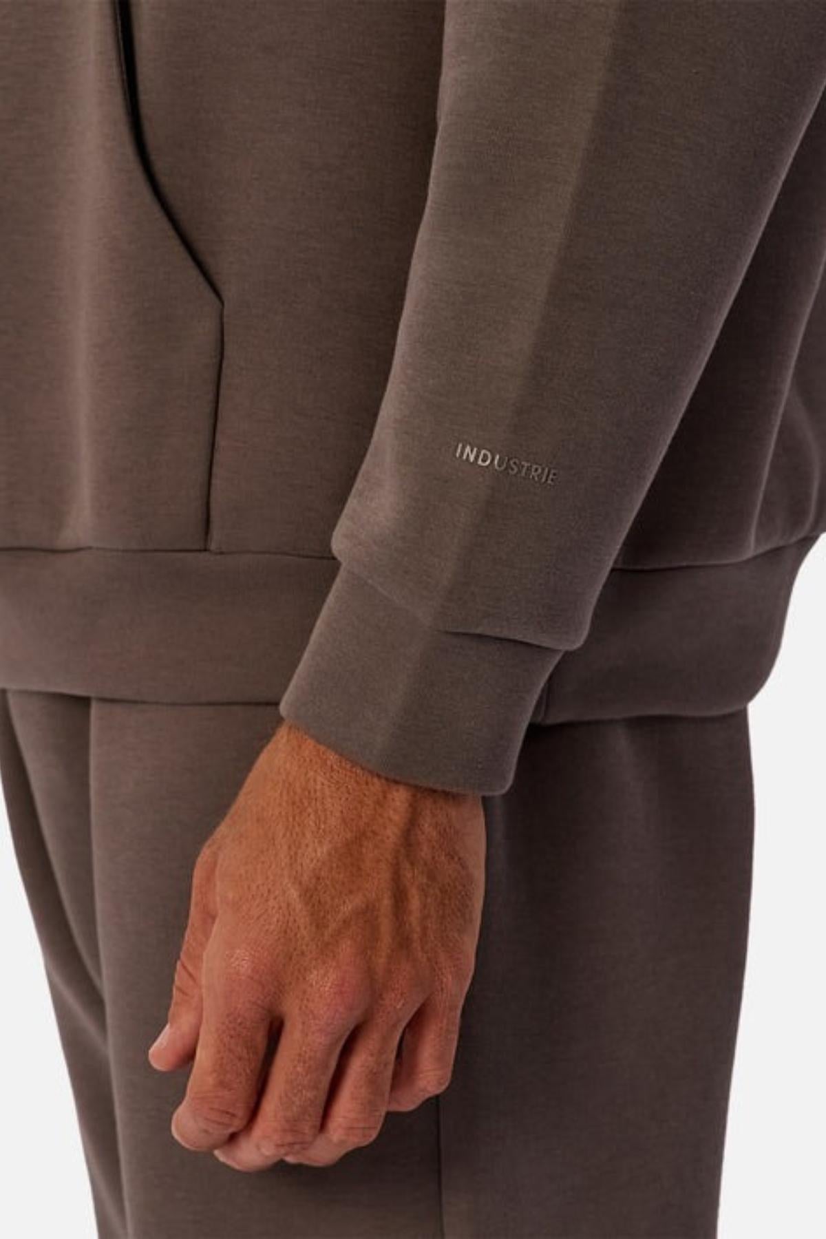 A close-up of a person in the Industrie The Tech Del Sur Hood, a brown men's pullover with matching pants. Their left hand is by their side, and “INDUSTRIE” is printed near the wrist on the sleeve.