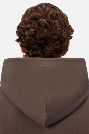 A person with curly brown hair is shown from behind, wearing the Industrie The Tech Del Sur Hood in brown with a small white INDUSTRIE logo on the back of the hood. The plain white background highlights its advanced athletic fabrication.