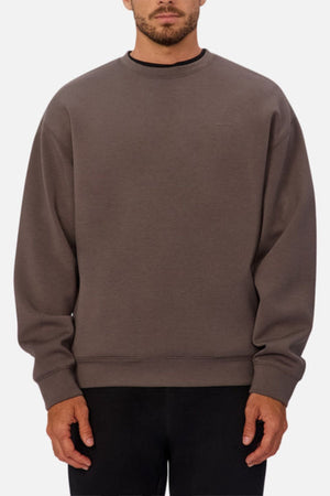 A man wears the Industrie The Tech Del Sur Sweat, a loose, long-sleeve dark gray crewneck with 4-way stretch and black pants. He stands against a plain light background, face partly visible, hands relaxed at his sides.