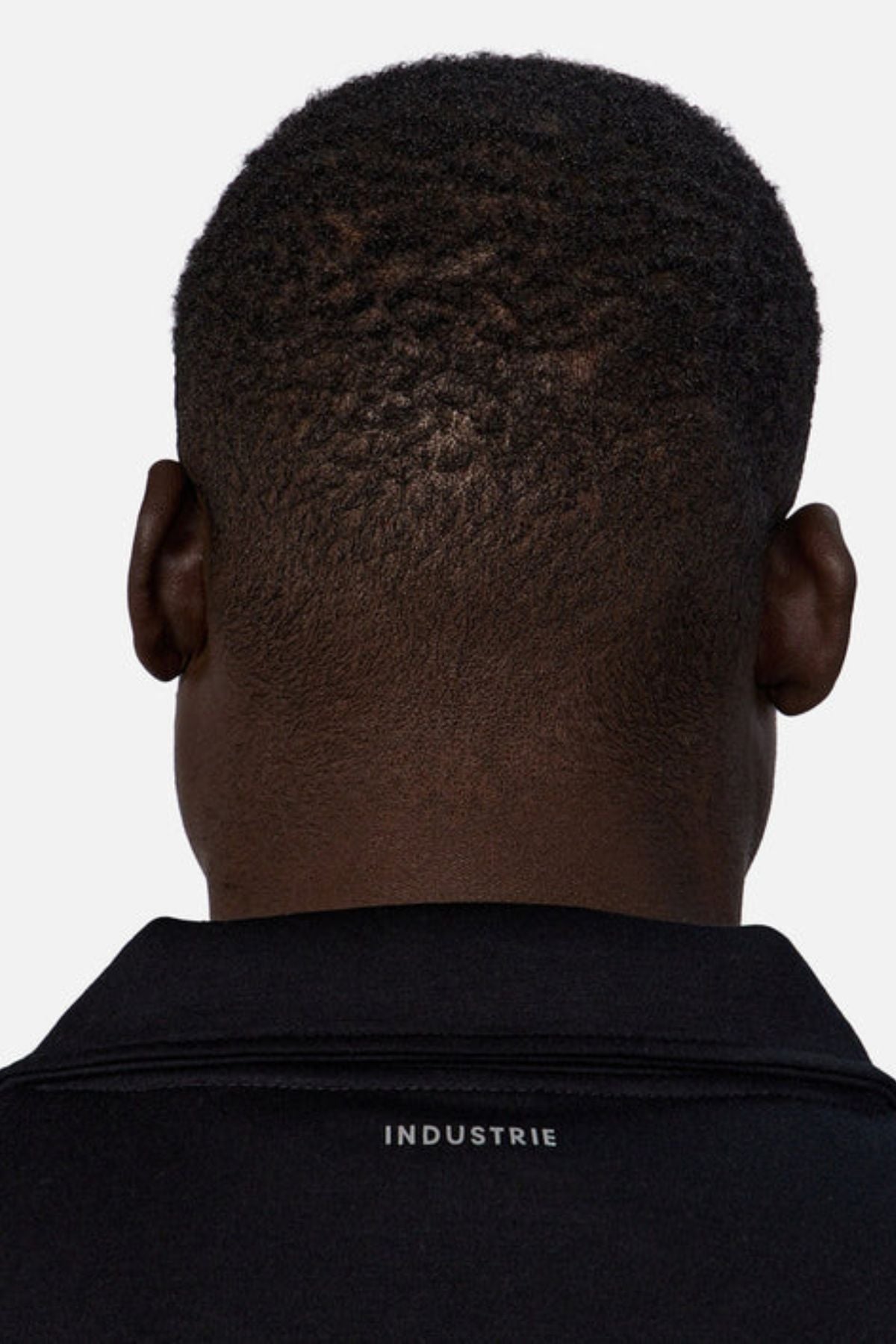A close-up of the back of a person’s head shows short, textured hair and a black Industrie The Tech Staunton Track Top with “INDUSTRIE” in white on the collar.