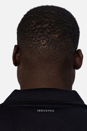A close-up of the back of a person’s head shows short, textured hair and a black Industrie The Tech Staunton Track Top with “INDUSTRIE” in white on the collar.