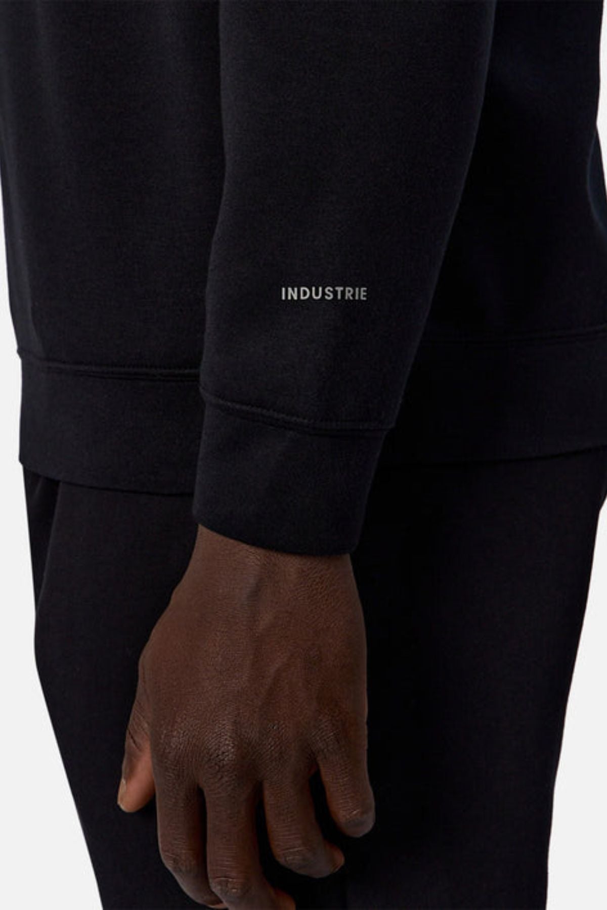 A close-up of a person’s arm and hand wearing the Industrie The Tech Staunton Track Top, featuring "INDUSTRIE" printed in white on the sleeve. The black athletic fabric contrasts with a plain, light background.