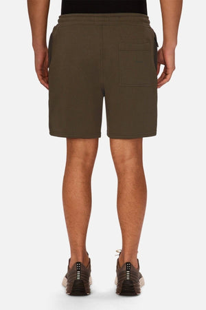 A person is standing facing away, wearing olive green Industrie The Tech Track Short, a black top, and gray sneakers. The athletic shorts feature a back pocket on the right side. The background is plain white.