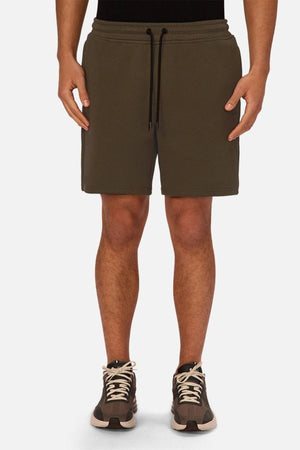 A person wearing dark olive green Industrie The Tech Track Short—a stylish take on men’s lounge shorts—and black sneakers with white laces, standing against a plain white background.
