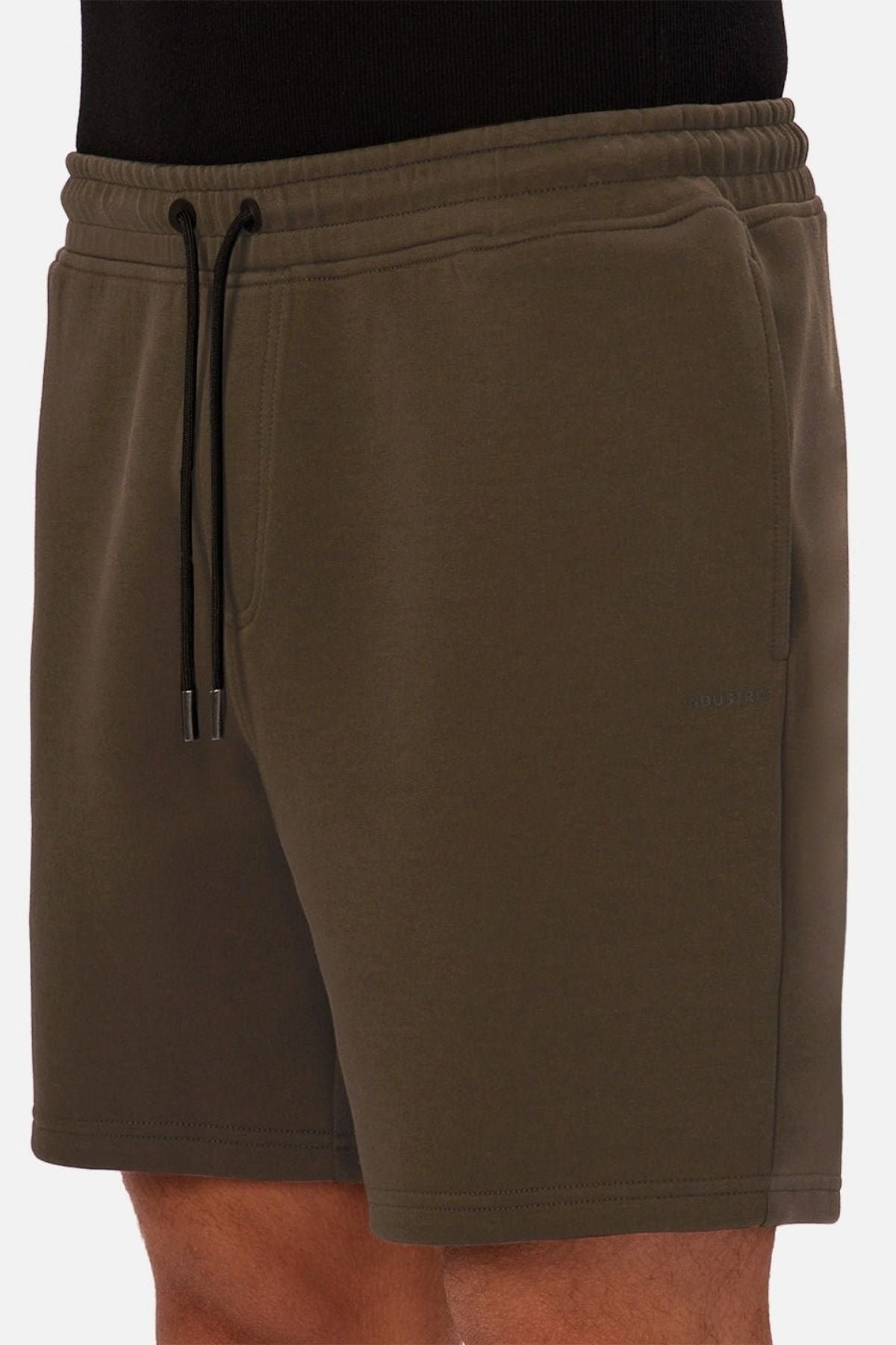 A person wearing Industrie The Tech Track Short in olive green, featuring side pockets, paired with a black shirt, shown from the waist to mid-thigh against a plain background.