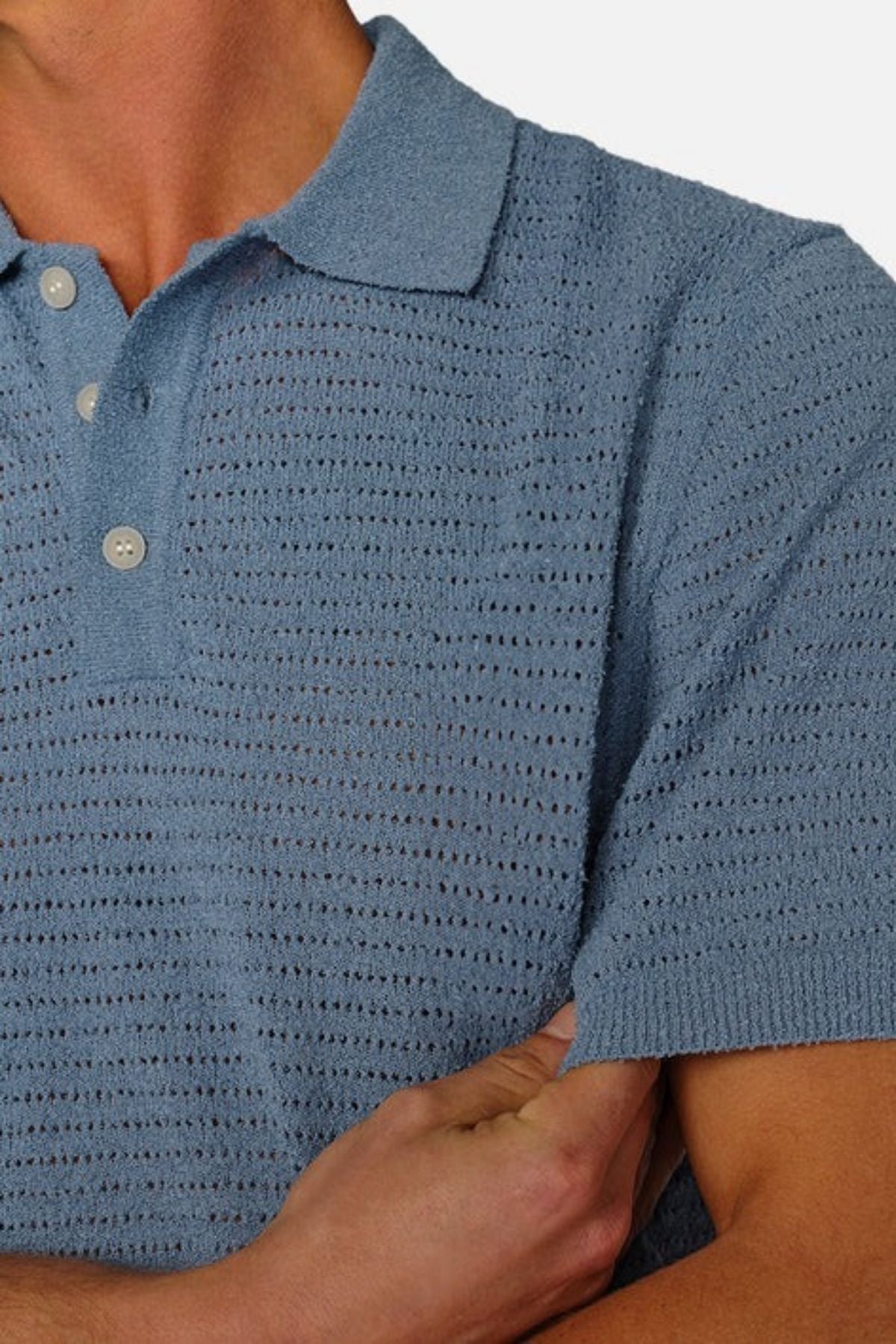 The individual wears a light blue Industrie The Terracina Polo with buttons, characterized by a vertical ribbed texture that imparts an airy feel. Their arms are crossed against a plain white background.
