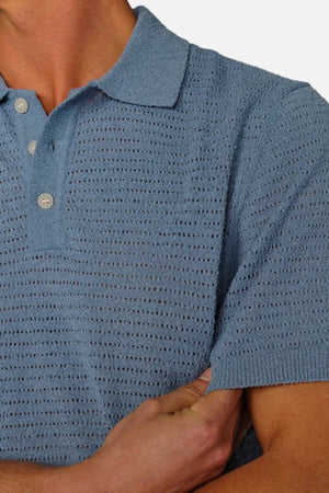 The individual wears a light blue Industrie The Terracina Polo with buttons, characterized by a vertical ribbed texture that imparts an airy feel. Their arms are crossed against a plain white background.