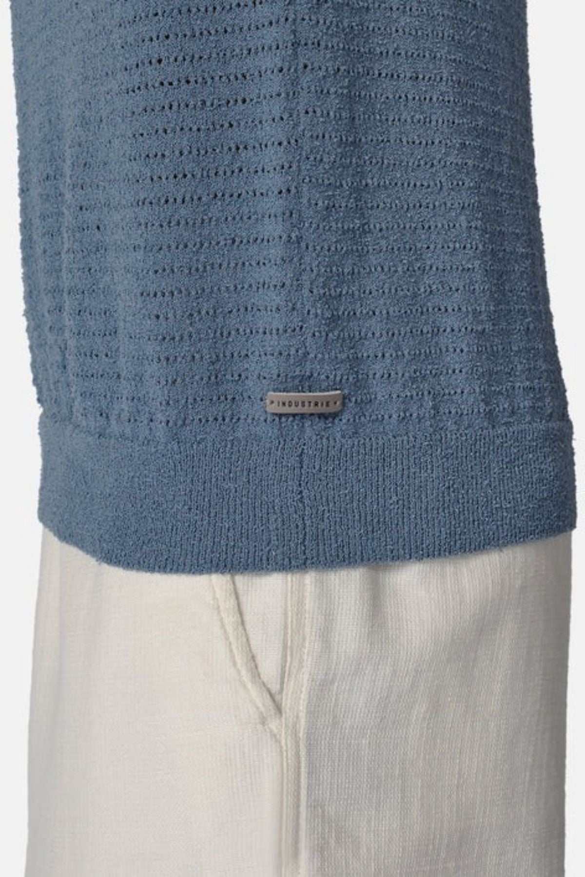 A close-up of a person wearing the Industrie The Terracina Polo, a textured blue sweater featuring vertical ribbed details, paired with off-white pants. Its lightweight material enhances comfort and includes a small rectangular label near the bottom.