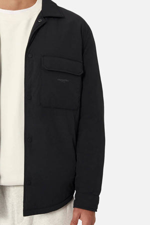 A person wearing the Industrie The Twin Cullman Jacket in black with quilted lining and a chest pocket over a white shirt and light pants, shown from shoulders to mid-thigh against a plain background. Subtle Industrie branding adds the finishing touch.