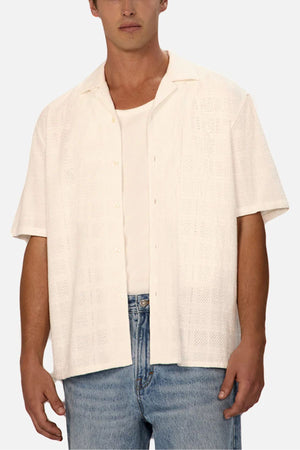 A man wears the Industrie The Venty Short Sleeve Shirt, an off-white, lightweight cotton shirt with a relaxed fit and subtle textured pattern, over a white tank top and light blue jeans against a plain backdrop.