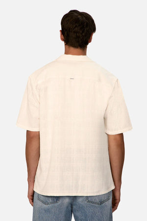 A person with short dark hair, seen from the back, wears an off-white Industrie The Venty Short Sleeve Shirt in relaxed fit and light blue jeans against a plain light background.