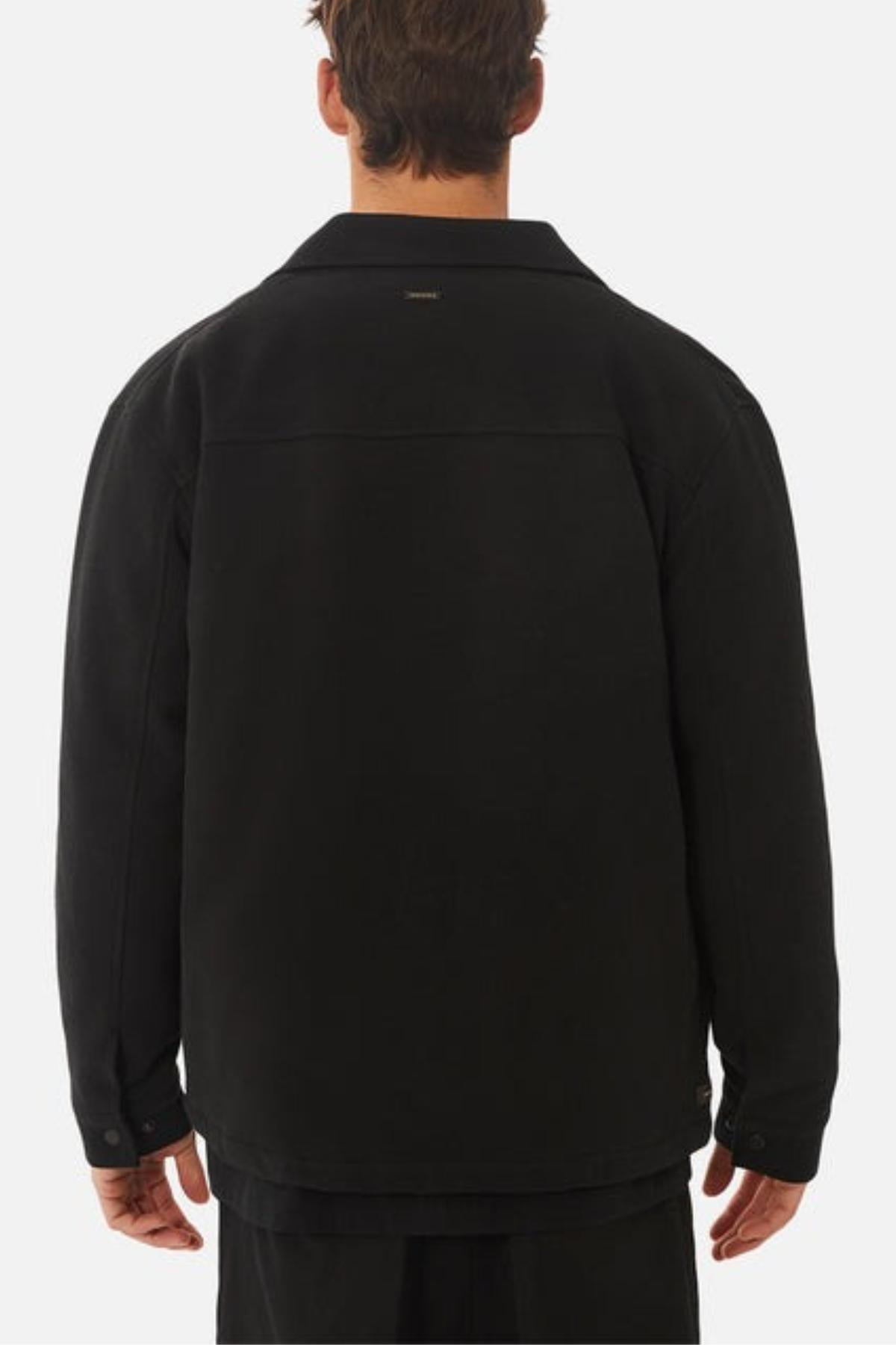 A man faces away from the camera, wearing the Industrie The Westport Jacket in black with long sleeves, a collar, and twin chest pockets, set against a plain light background.