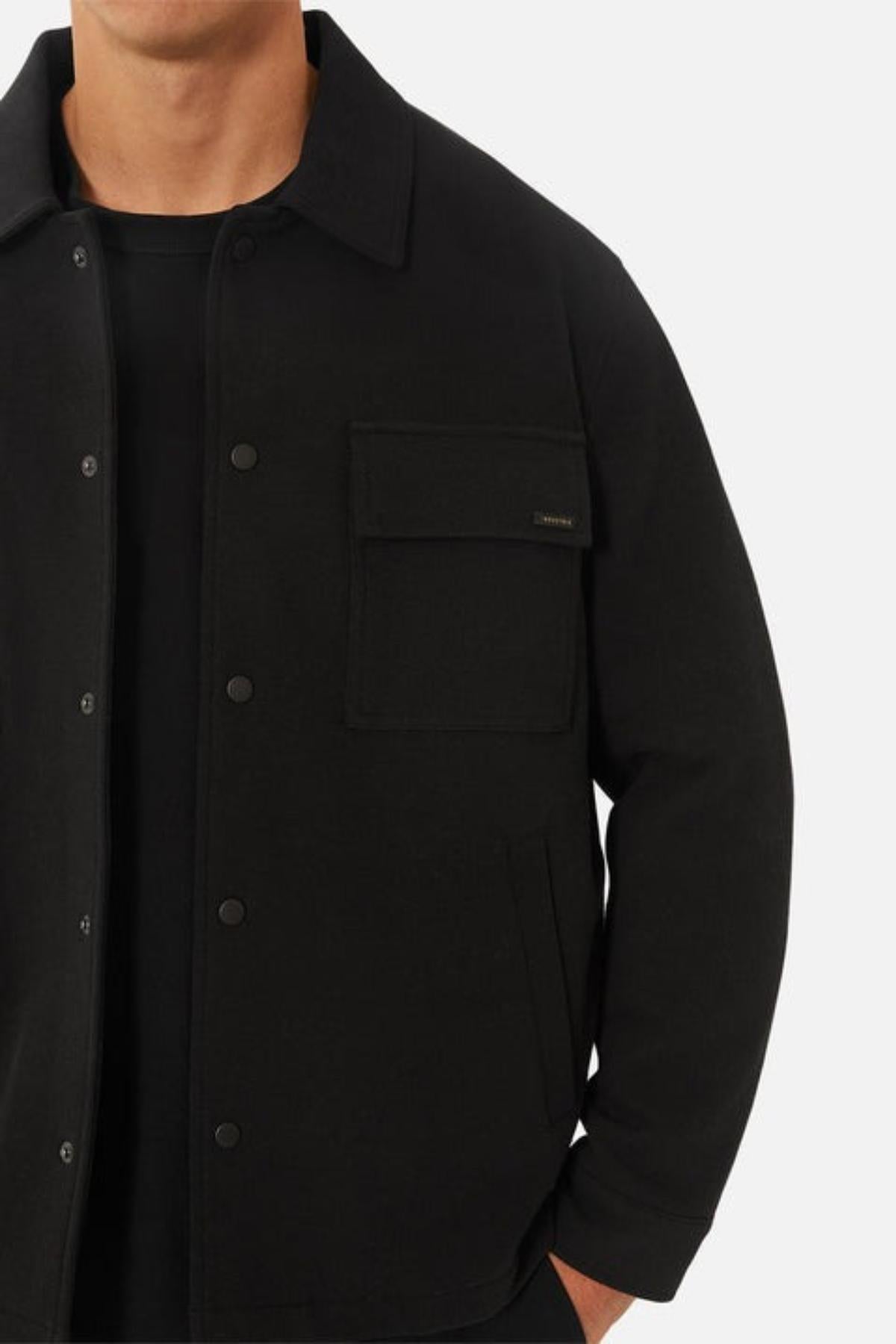 A man wearing the Industrie The Westport Jacket in black, featuring a large chest pocket, side pockets, and quilted lining, stands against a plain light background with his face not visible.