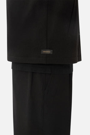 A close-up of a person wearing black pants and the Industrie The Westport Jacket in black, with twin chest pockets and a small "INNVIERTE" label near the hem. The plain, light background highlights the jacket’s clean lines.