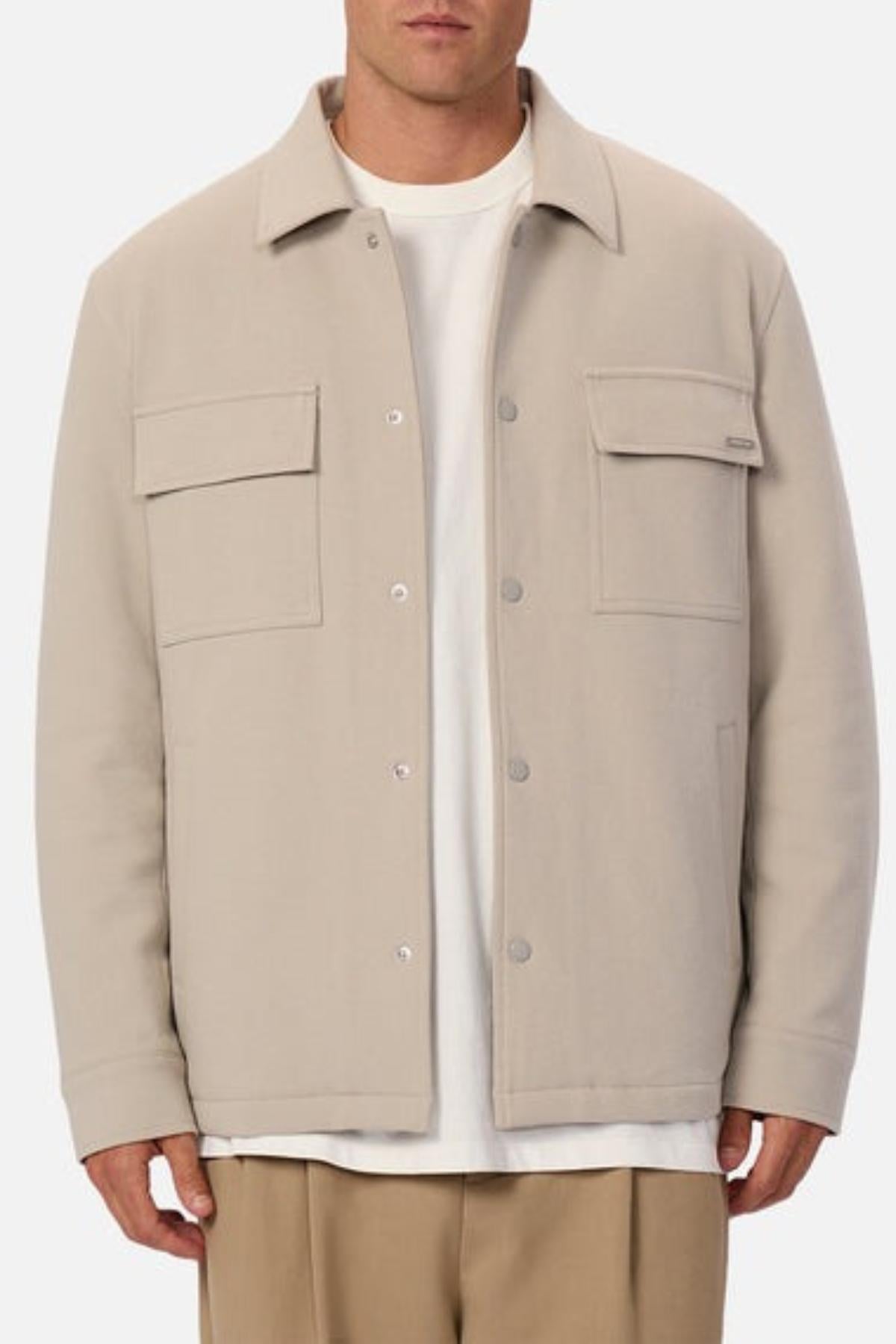 Someone stands against a plain background wearing the Industrie The Westport Jacket in light beige, featuring twin chest pockets and two front flap pockets, layered over a white t-shirt and tan trousers.