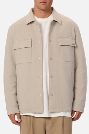 Someone stands against a plain background wearing the Industrie The Westport Jacket in light beige, featuring twin chest pockets and two front flap pockets, layered over a white t-shirt and tan trousers.