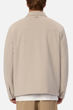 A person is seen from behind wearing the Industrie The Westport Jacket in light beige, featuring twin chest pockets, over a white shirt and beige pants against a plain, light-colored background.