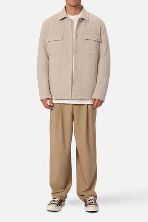 A man faces forward against a white background, wearing an Industrie The Westport Jacket in light beige with twin chest pockets over a white shirt, loose tan pants, and beige low-top sneakers featuring white laces.