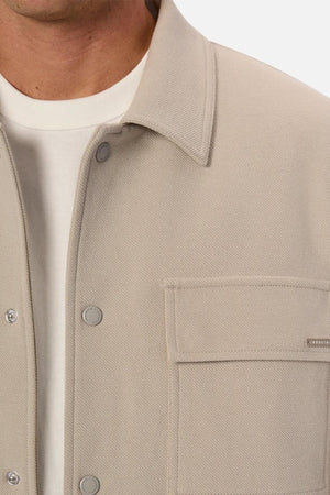 Close-up of a person wearing the Industrie The Westport Jacket in light beige with snap buttons over a white T-shirt, highlighting part of the chest, collar, and one textured twin chest pocket.