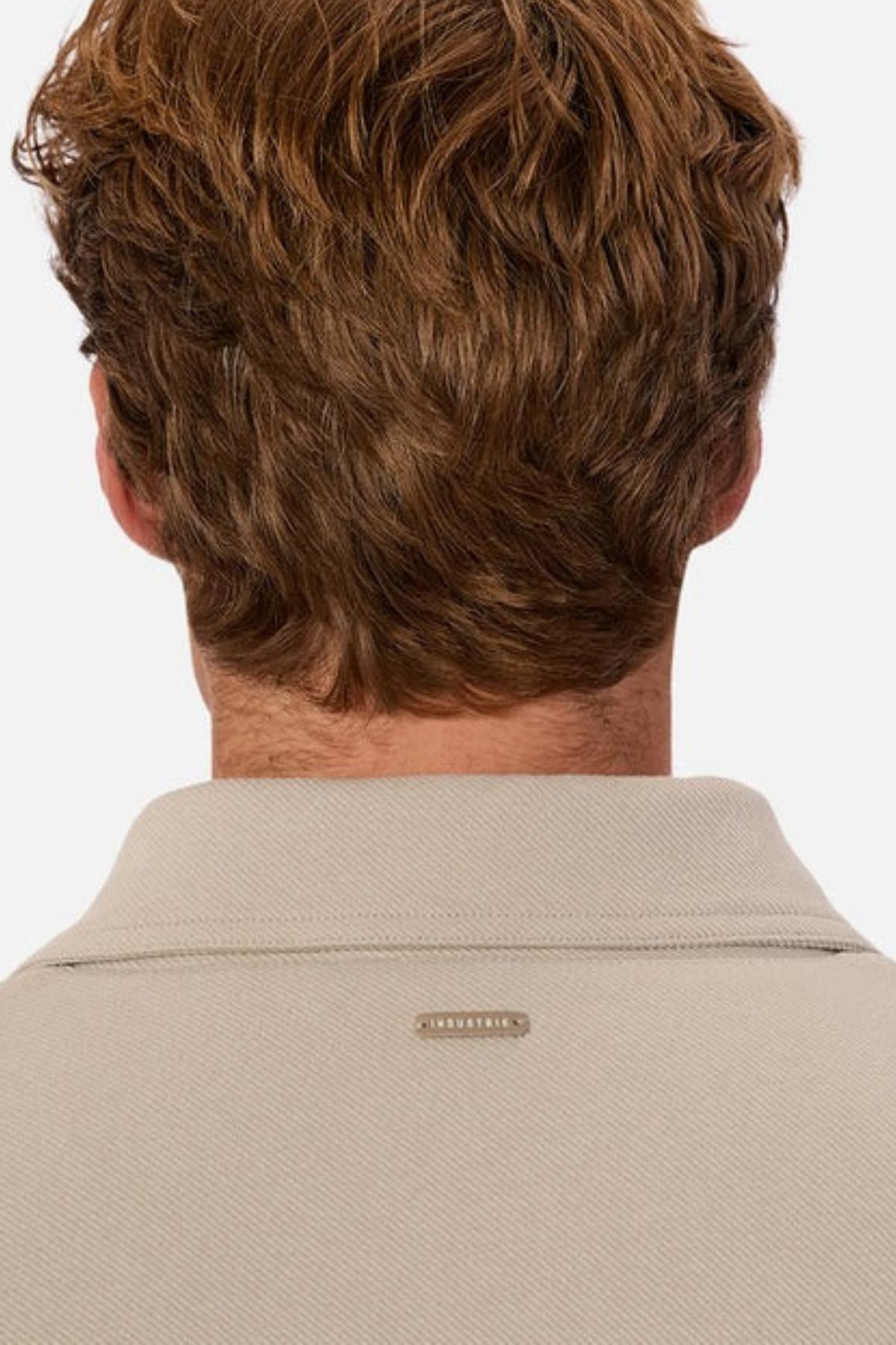 A person with short, wavy, light brown hair is shown from behind, wearing the beige Industrie The Westport Jacket featuring a small rectangular metal tag on the upper collar and twin chest pockets for classic style.
