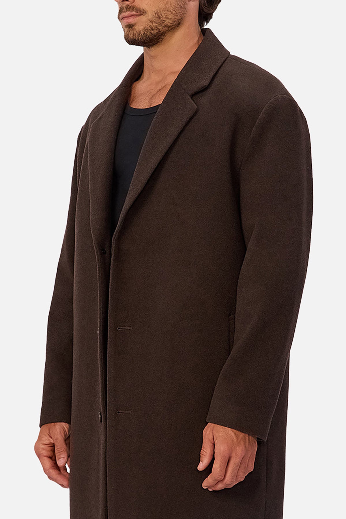 A man wears the Industrie The Wool Blend Jacques Coat, a knee-length dark brown wool blend coat with a structured fit, over a black shirt, standing against a plain white background; his face is partially out of frame.