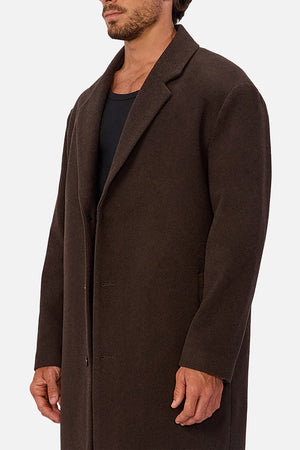 A man wears the Industrie The Wool Blend Jacques Coat, a knee-length dark brown wool blend coat with a structured fit, over a black shirt, standing against a plain white background; his face is partially out of frame.