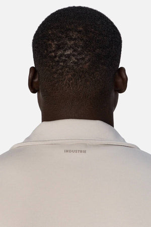 A person with short, curly black hair is seen from behind wearing the Industrie The Zip Thru Track Top, a light-colored collared shirt with INDUSTRIE branding on the back collar. The background is plain and light gray.