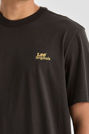 A person is shown from the upper torso up, wearing a relaxed fit Lee Originals Relaxed Tee in plain black with short sleeves and a small yellow "Lee® Originals" logo on the left chest. The T-shirt is made from 100% cotton.