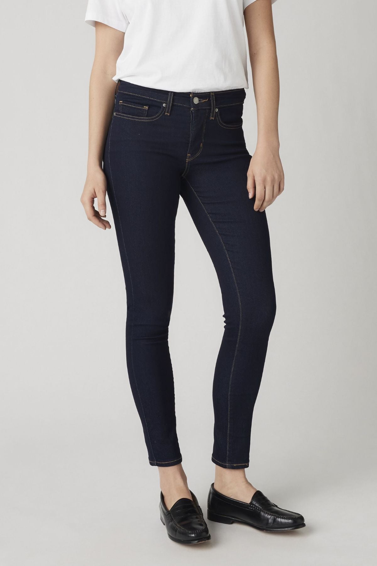 A person wearing a white t-shirt, Levis 311 Shaping Skinny Jean in dark blue, and black loafers stands against a plain light background. Their face is not visible in the image.