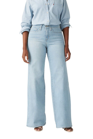 Wearing Levis 318 Shaping Wide Leg Jeans in light blue and a matching light blue button-up shirt with rolled sleeves, this person stands against a white background, showing a flattering silhouette from shoulders to feet.