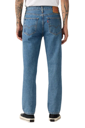 A person wearing Levis 516 Straight Jeans in light blue stonewash, a white shirt, and black sneakers is shown from the back. The comfy jeans have two back pockets and a brown leather patch. Their arms display tattoos.