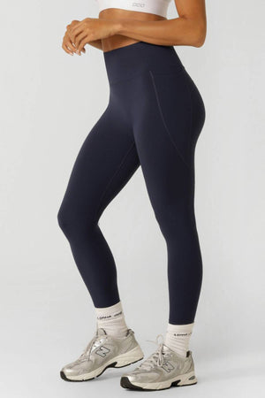 A person wearing a white sports bra, white socks, gray sneakers, and high-waisted Lorna Jane Amy No Ride Phone Pocket Ankle Biter Leggings in navy stands against a plain light background. Their body is shown from shoulders to feet.