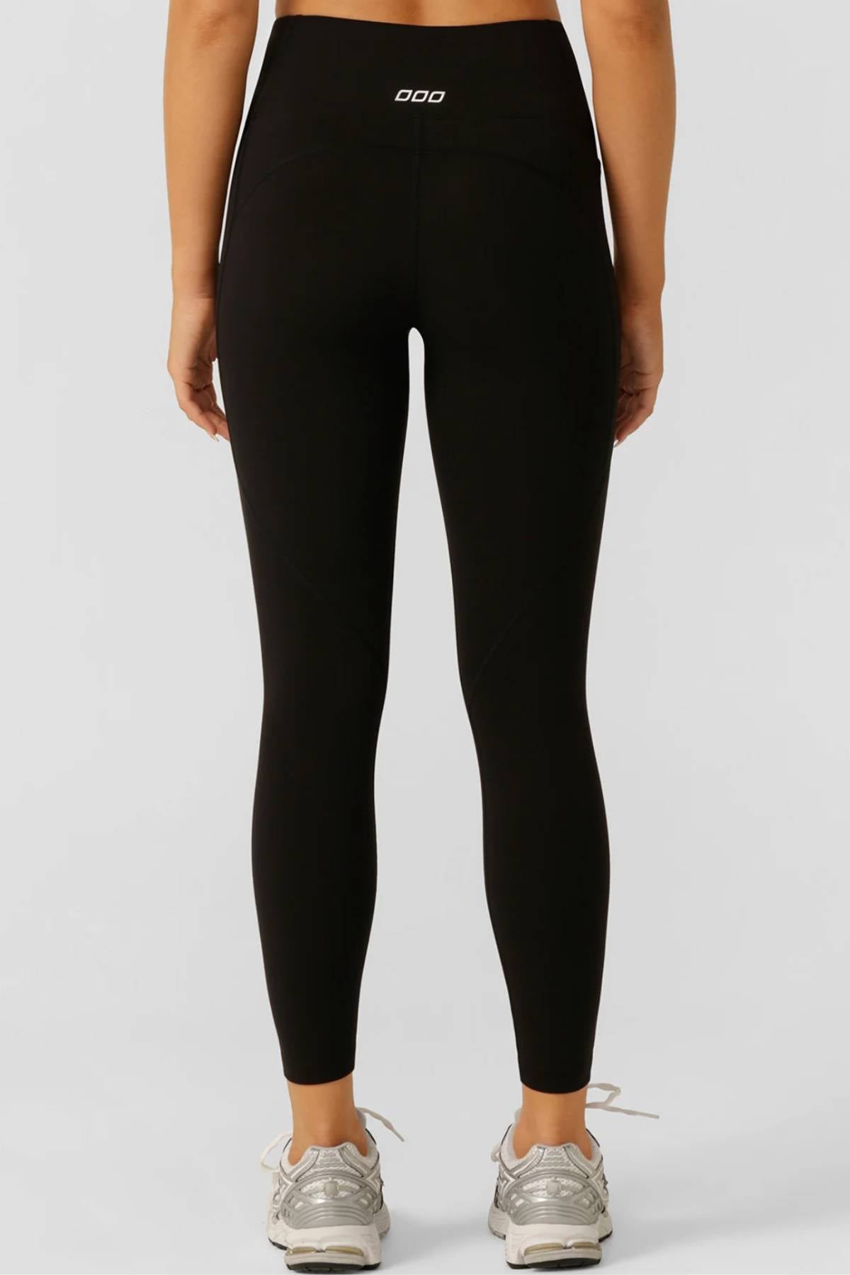 A person faces away, wearing black Lorna Jane Amy No Ride Phone Pocket Ankle Biter Leggings with a white logo on the waistband and light gray sneakers. The plain light background highlights the leggings’ sleek workout style.
