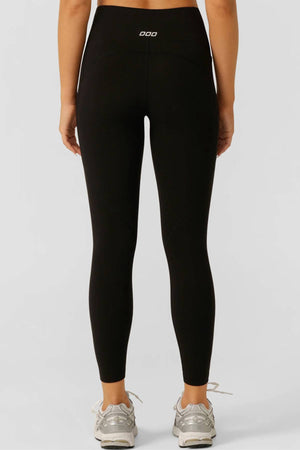 A person faces away, wearing black Lorna Jane Amy No Ride Phone Pocket Ankle Biter Leggings with a white logo on the waistband and light gray sneakers. The plain light background highlights the leggings’ sleek workout style.