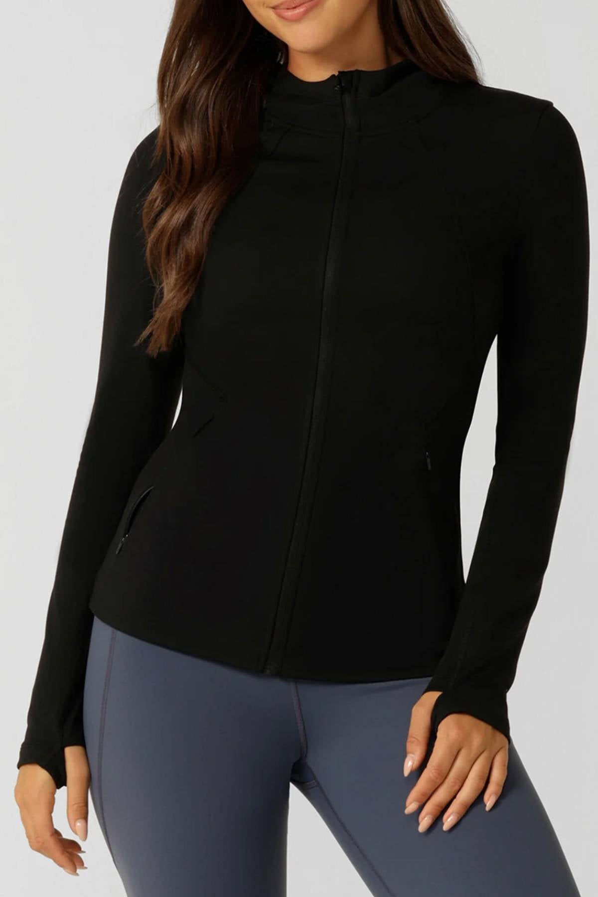 Woman wearing the Lorna Jane Amy Ultimate Thermal Hooded Jacket—black, fitted, with long sleeves and thumbholes—paired with high-waisted gray leggings. Her long brown hair is loose and wavy against a plain light background. Perfect for winter workouts.