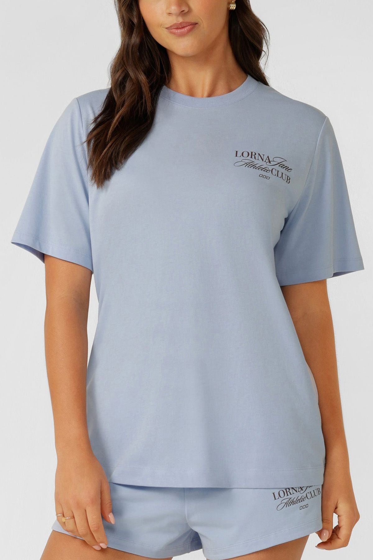 A woman wears a light blue Lorna Jane Athletic Club Relaxed T-Shirt and matching shorts, both featuring "Lorna Jane Fitness Club" text, made from a breathable cotton blend. She stands against a plain background.