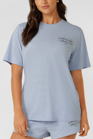 A woman wears a light blue Lorna Jane Athletic Club Relaxed T-Shirt and matching shorts, both featuring "Lorna Jane Fitness Club" text, made from a breathable cotton blend. She stands against a plain background.