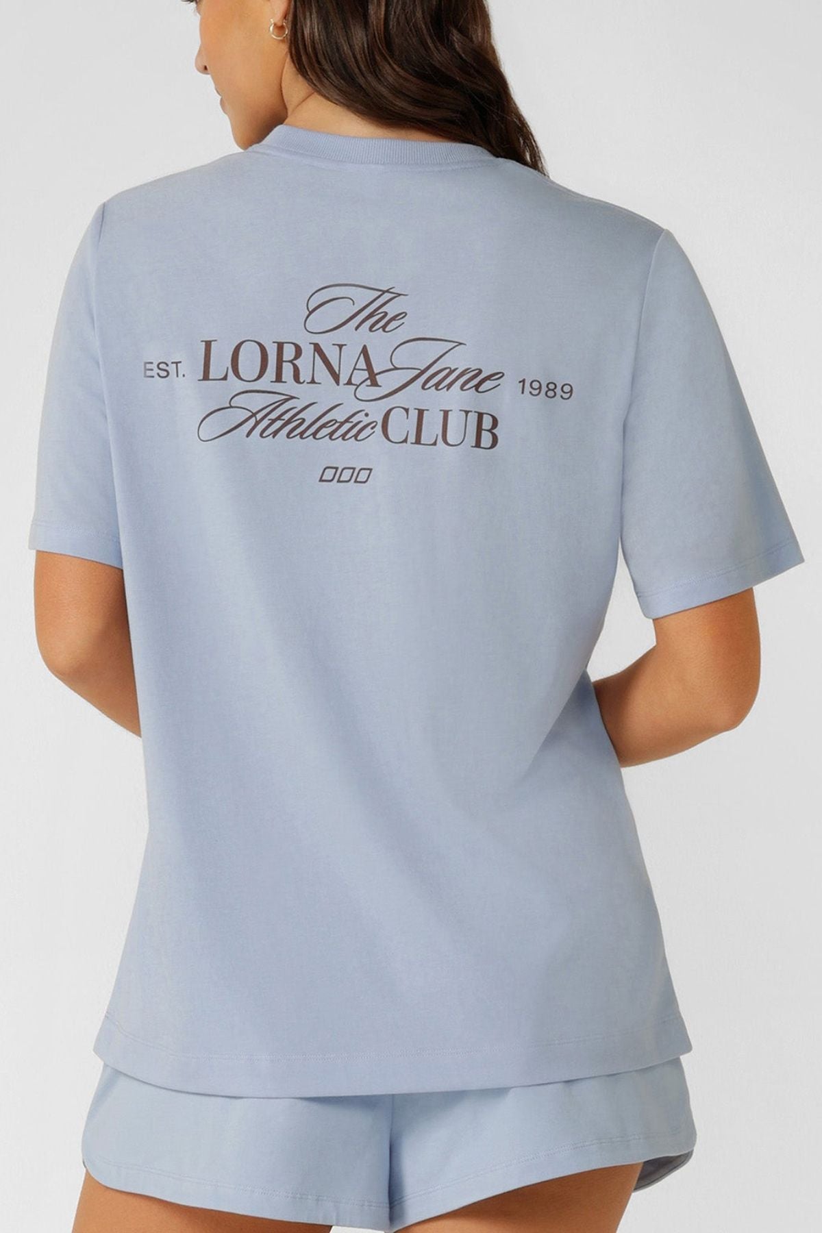 A woman is shown from the back wearing a relaxed fit, light blue Lorna Jane Athletic Club Relaxed T-Shirt and matching shorts. Made from a breathable cotton blend, the tee has "The Lorna Jane Athletic Club EST. 1989" in black script on the back.