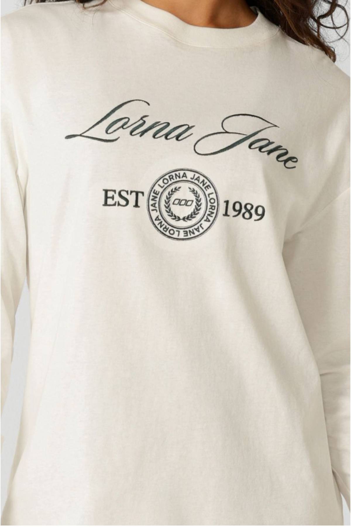 A person wears the Lorna Jane Country Club Long Sleeve T-Shirt, made of 100% cotton, featuring "Lorna Jane" in cursive, "EST 1989," and a round Lorna Jane logo on the front.
